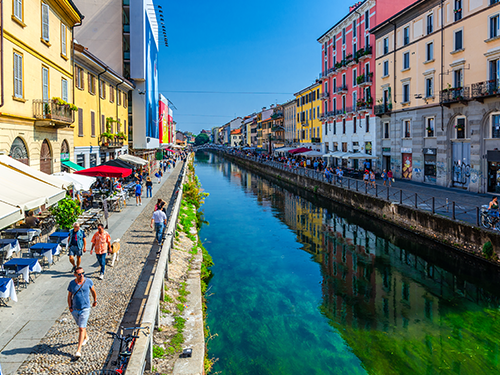 Milan  pictured on a sunny day, with people walking down the canal in the Navigli area 