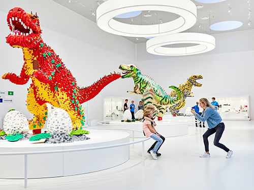 A woman takes a picture of two children who are sat in front of a large lego dinosaur display 