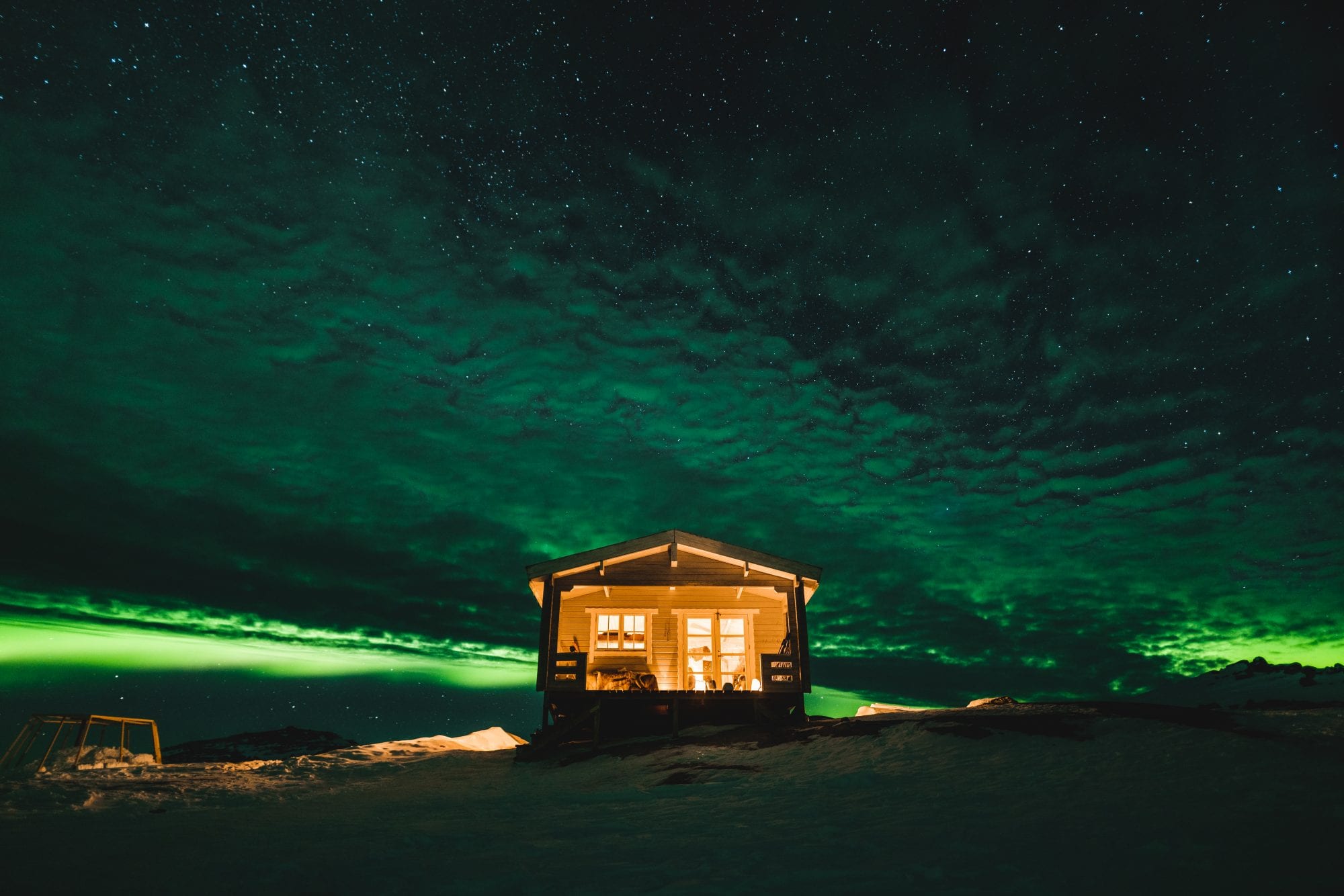 A cosy cabin in the wintery Icelandic landscape lit up inside and with Northern Lights shining overhead