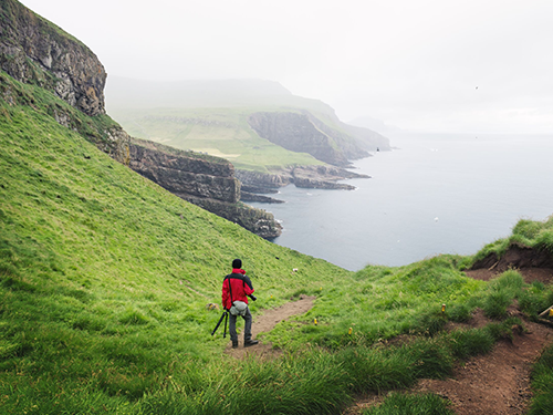 A man in a red rain jacket is pictured holding photography equipment as he looks out over a view of the cliffs of the Faroe Islands