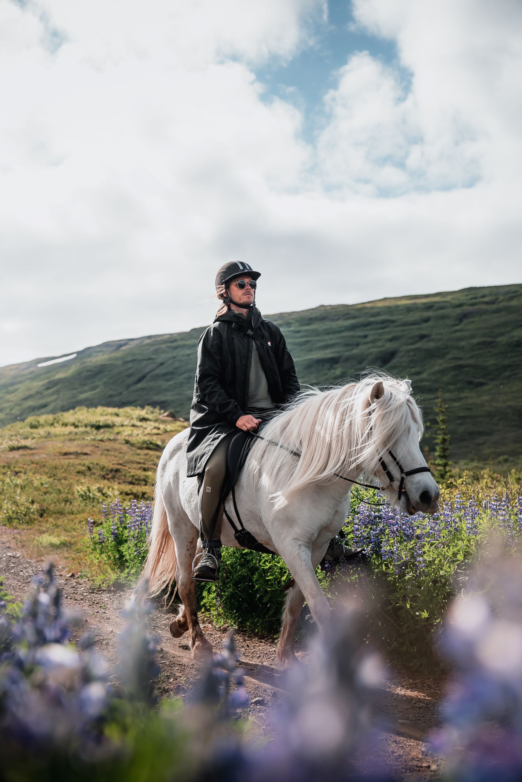 Rurik Gislason sits on a white Icelandic horse, riding a trail with lupin flowers in the foreground