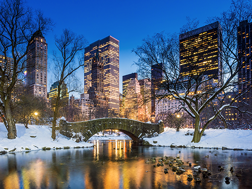 The Woolman Park lit up in an evening glow with the city lights reflecting on the water in the pond and snow laying on either side of the bridge