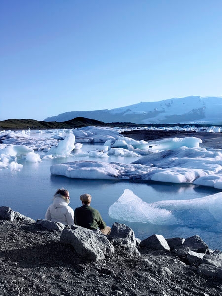 Image of a man and a woman sitting on the sandy beach by the Glacial lagoon in the south coast of Iceland.