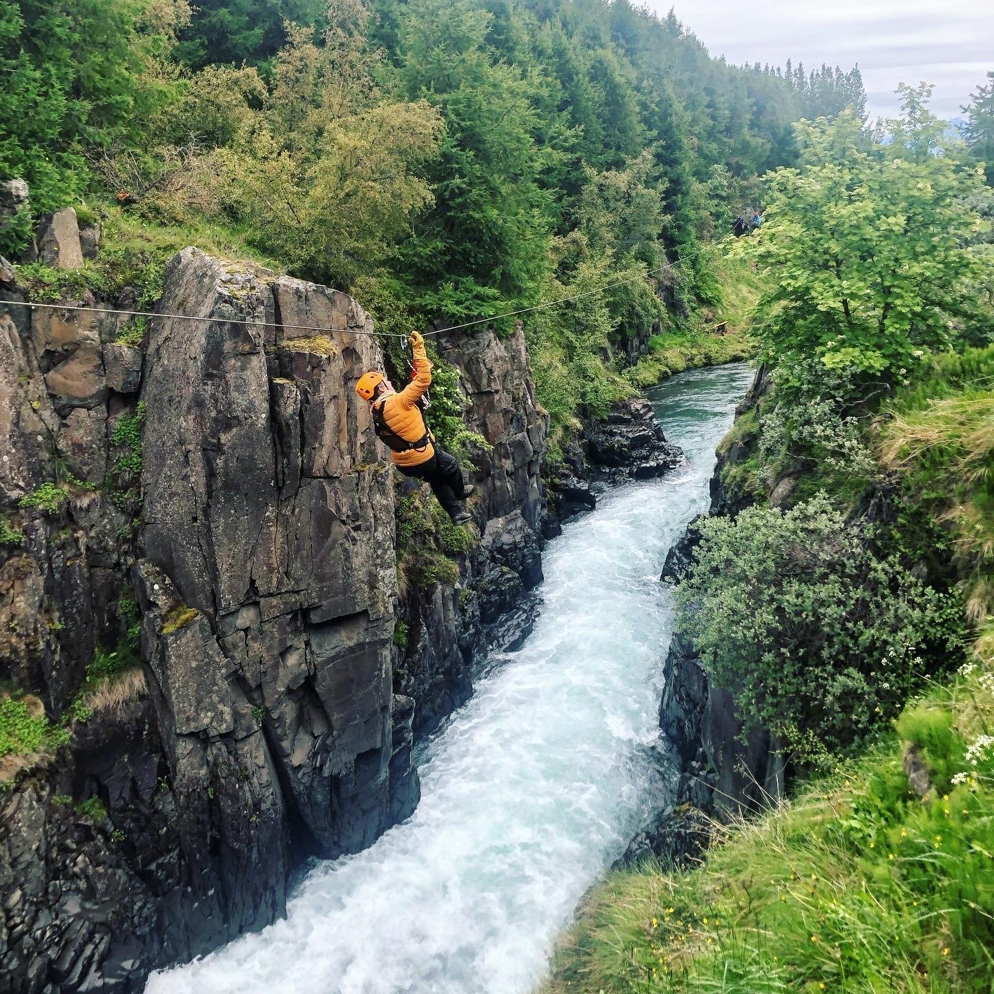A person is harnessed to a zipline and is traveling high above river rapids, with a rocky canyon and trees on either side
