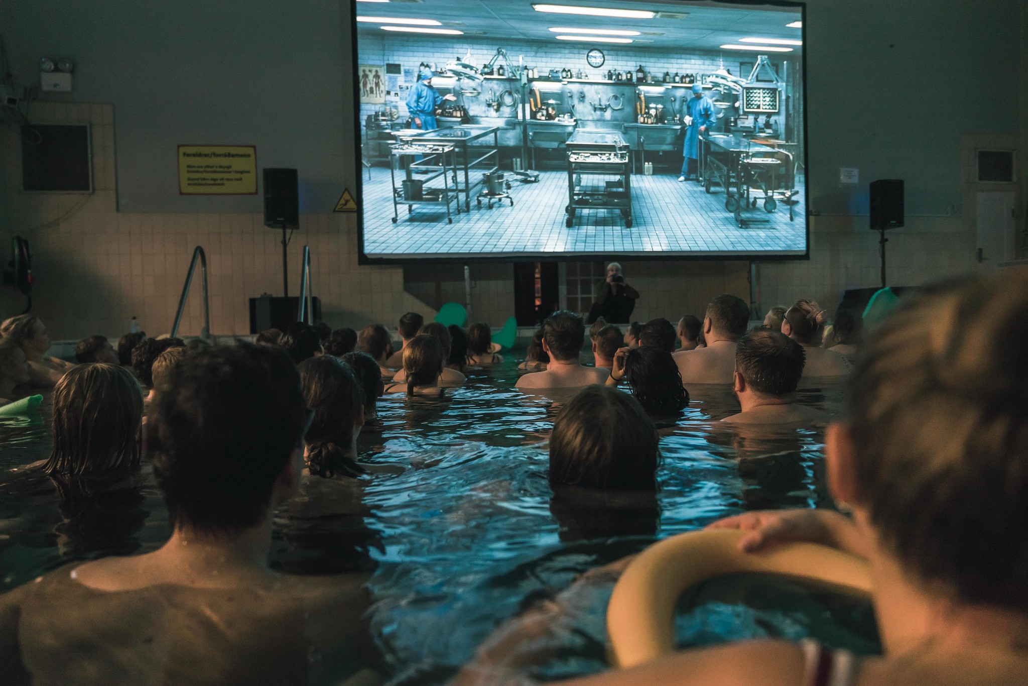 A group of people bathing in a public pool with a screen overhead that is playing a film as part of Riff International Film Festival