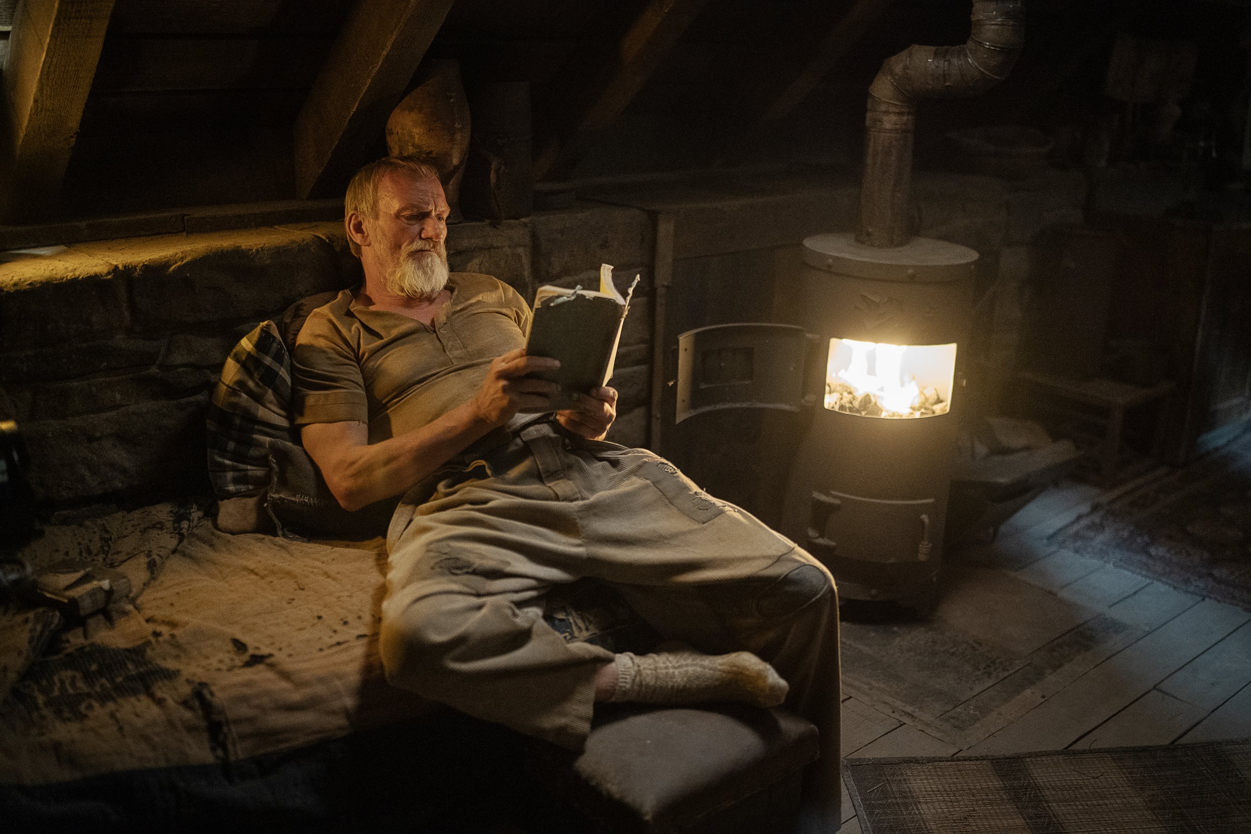 Image of Ingvar Sigurðsson reclining on an old sofa, reading by an open fire. A scene from Rebel Moon TV series
