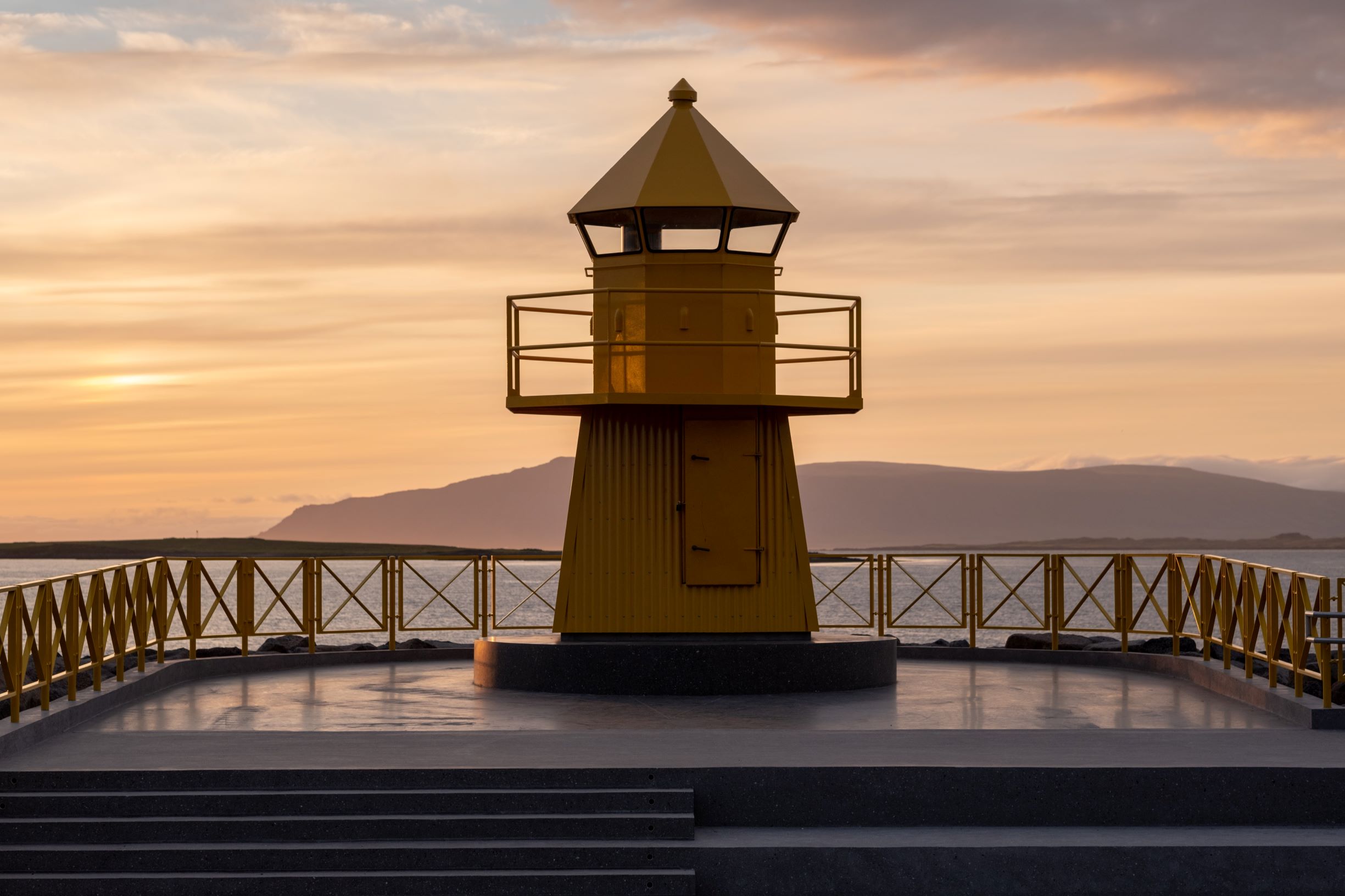 A yellow lighthouse shaped structure positioned by the shore in Reykjavik, with a background of Esjan mountain