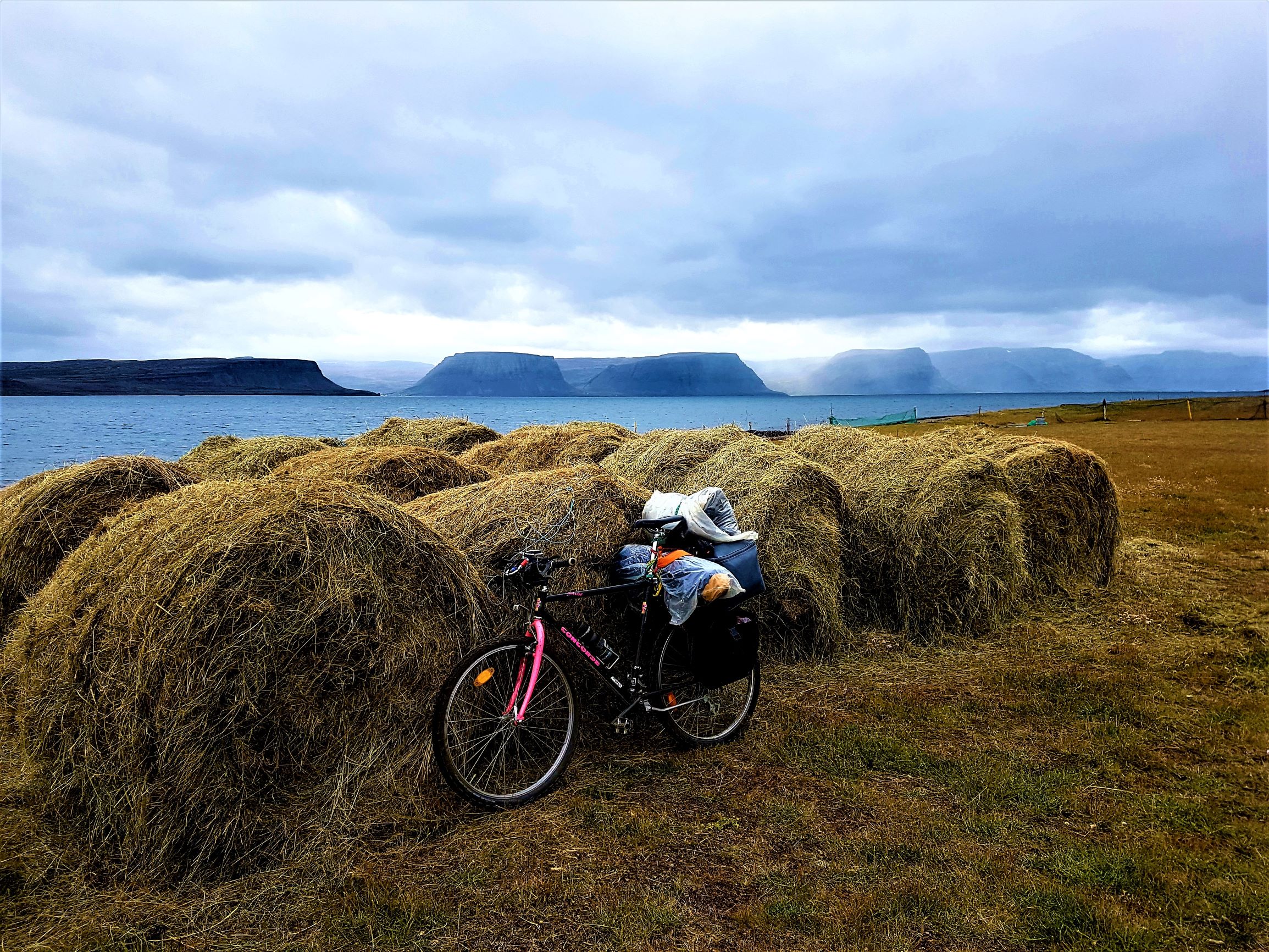 A bike with bags on the back leaning against a hay bale with a view of the Westfjords of Iceland in the background