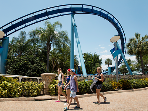 A family group walk through a theme park, with a blue rollercoaster pictured in the background 