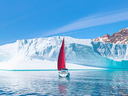 A sail boat with a red sail is pictured with a large ice berg in the background 