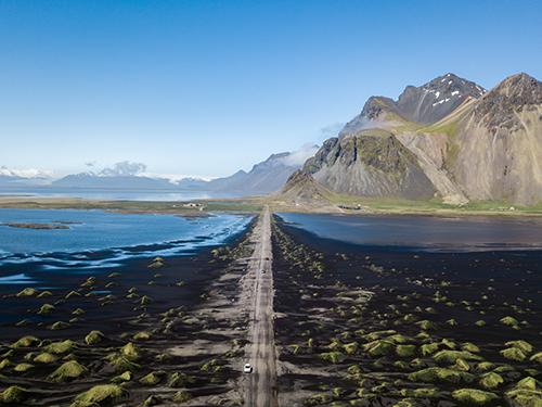 A view of the roads of the South East of Iceland, with a singular white care making it’s way past a mountain on a blue sky day 