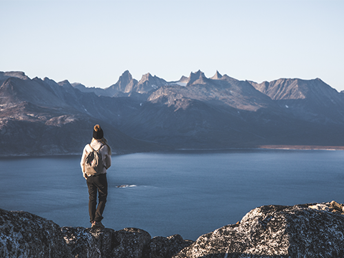 A person stands on a rocky surface in South Greenland with their back to the camera. In front of them is a body of water and a mountainous view 