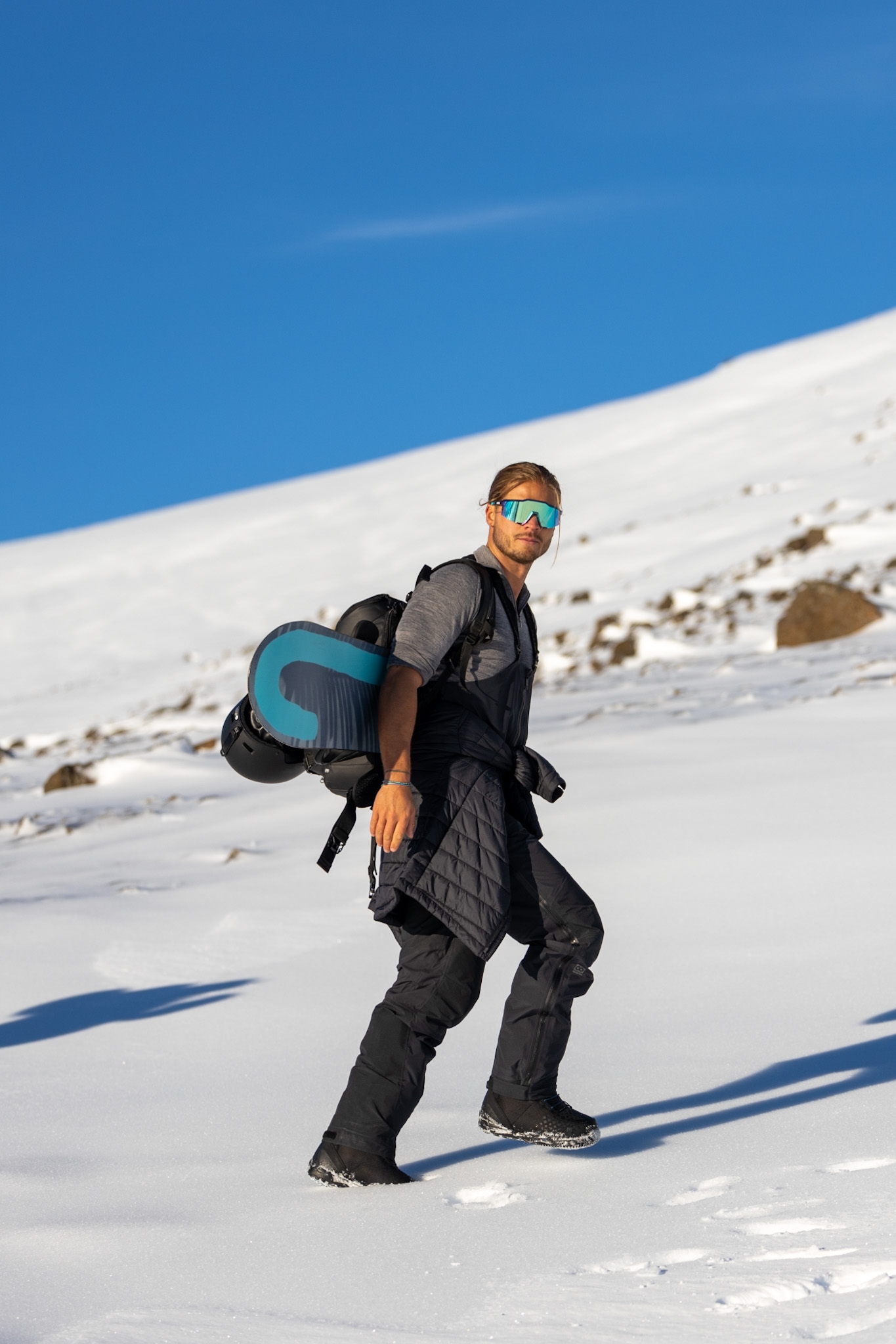 Rúrik Gíslason in ski gear carries a snowboard on his back and stands on a sno-covered hillside with blue skies overhead