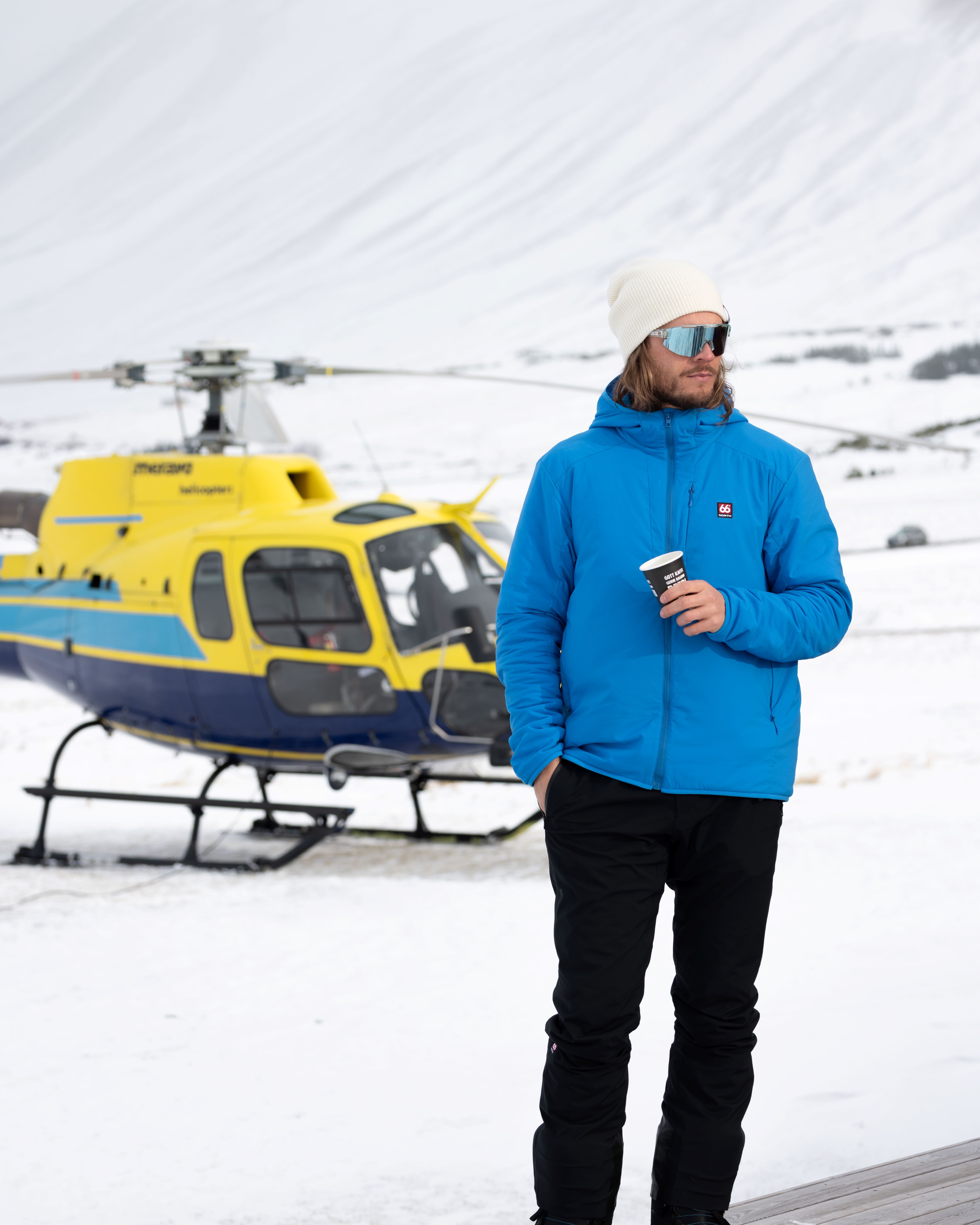 Rúrik Gíslason stands on snowy ground with a backdrop of snow-covered mountains and a helicopter used for heli-skiing adventures in North Iceland.