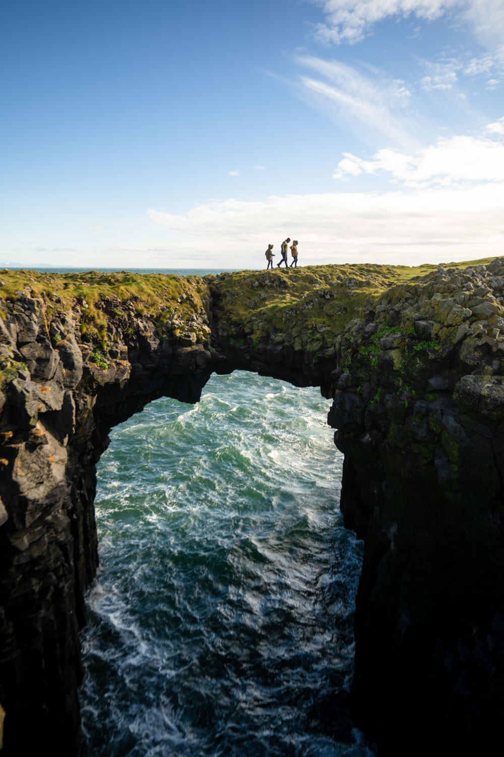 Chris Burkard and his 2 sons walk across a rocky archway at Arnastapi on the Snæfellsnes peninsula. The sea is underneath the arch