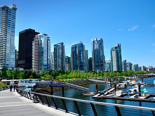 A cityscape of Vancouver as pictured from the harbour area, where several boats are docked and people are mulling around 