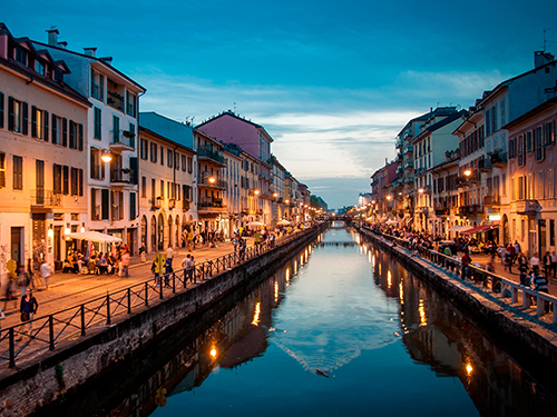 An evening scene of the water by the canal at Navigli, Milan, with people milling around in eateries and bars 