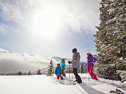 A family of four stand at the top of a ski slope in Denver, holding hiking poles and looking out over the stunning vistas 