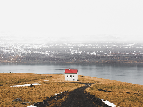 A white and red isolated house pictured from the top of the road leading up to it, with a foggy Westfjords scene behind