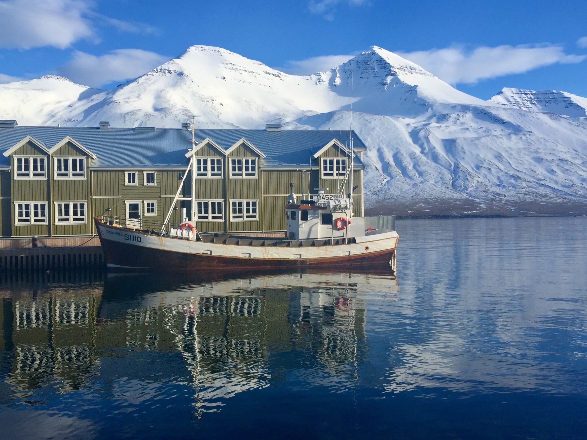 A boat is moored outside a green waterfront building in Siglufjörður, with snowy mountains as a backdrop