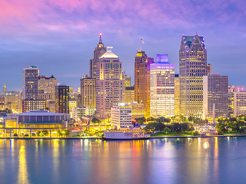 A city scape of Detroit as viewed from across the water, with the bright city lights lit up against a dusky evening sky 