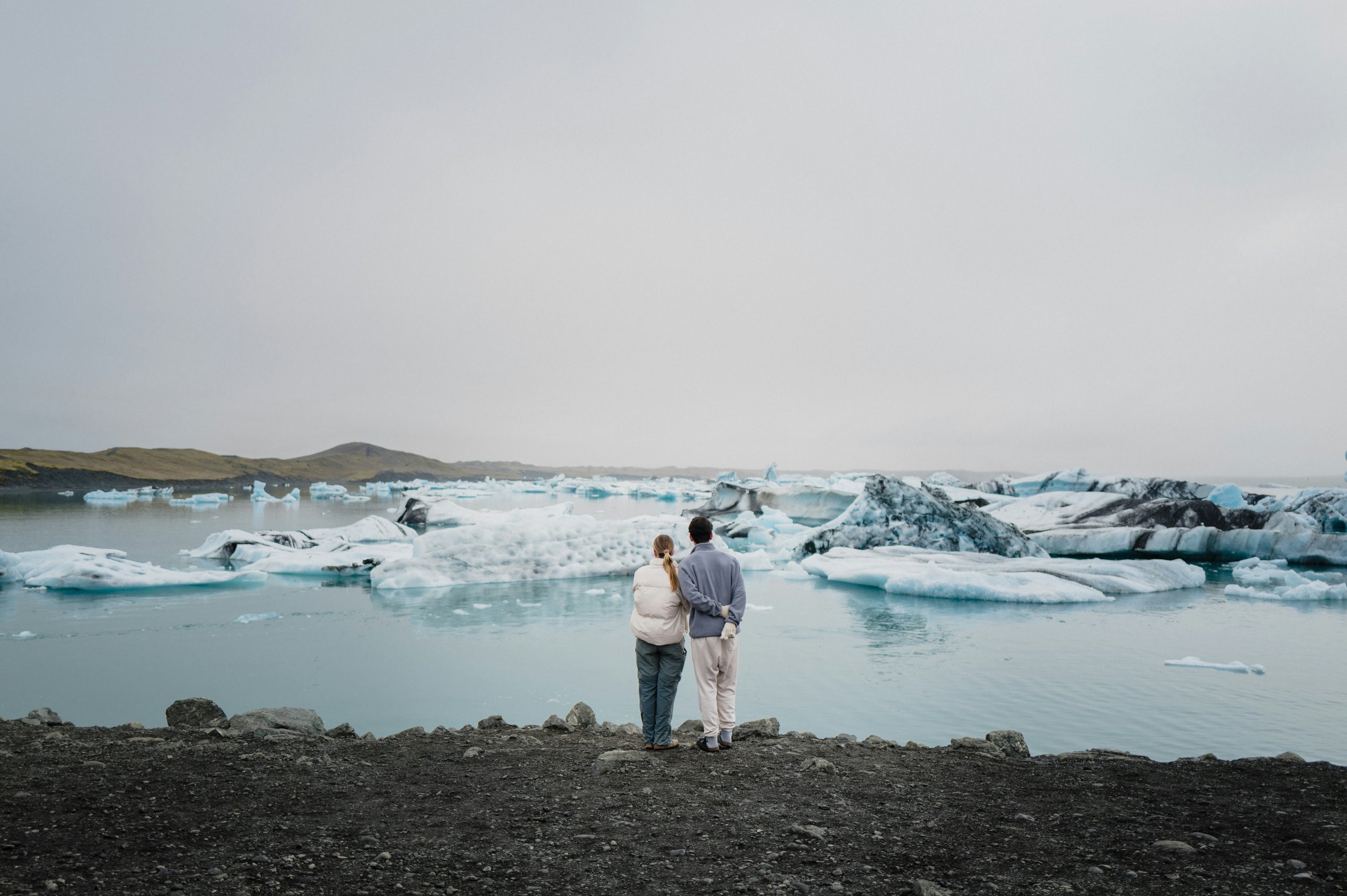 a couple stand next to each other, their backs to the camera, looking out to a view of Jökulsárlón glacier lagoon