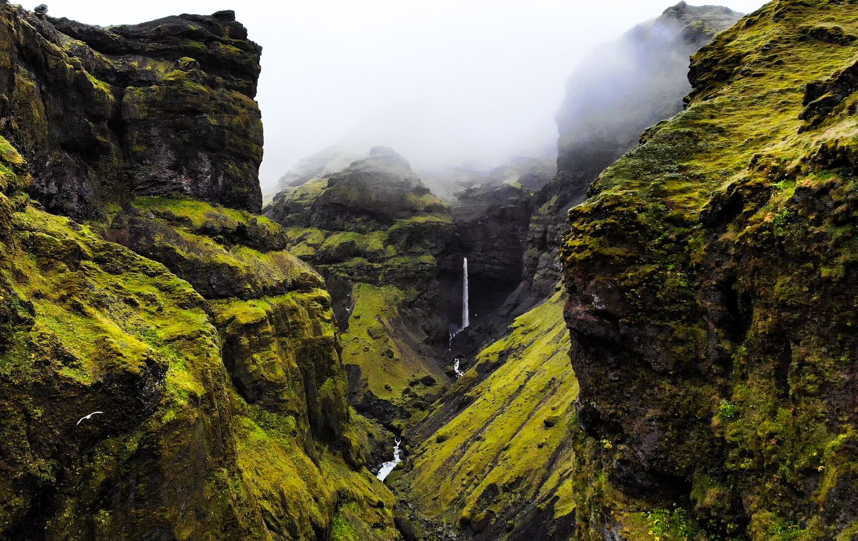 Views of mountains in the clouds in Vatnajökull National Park. The picture shows Mulagljufur valley, a lush green, mountainous landscape with a small waterfall in the centre of the picture.