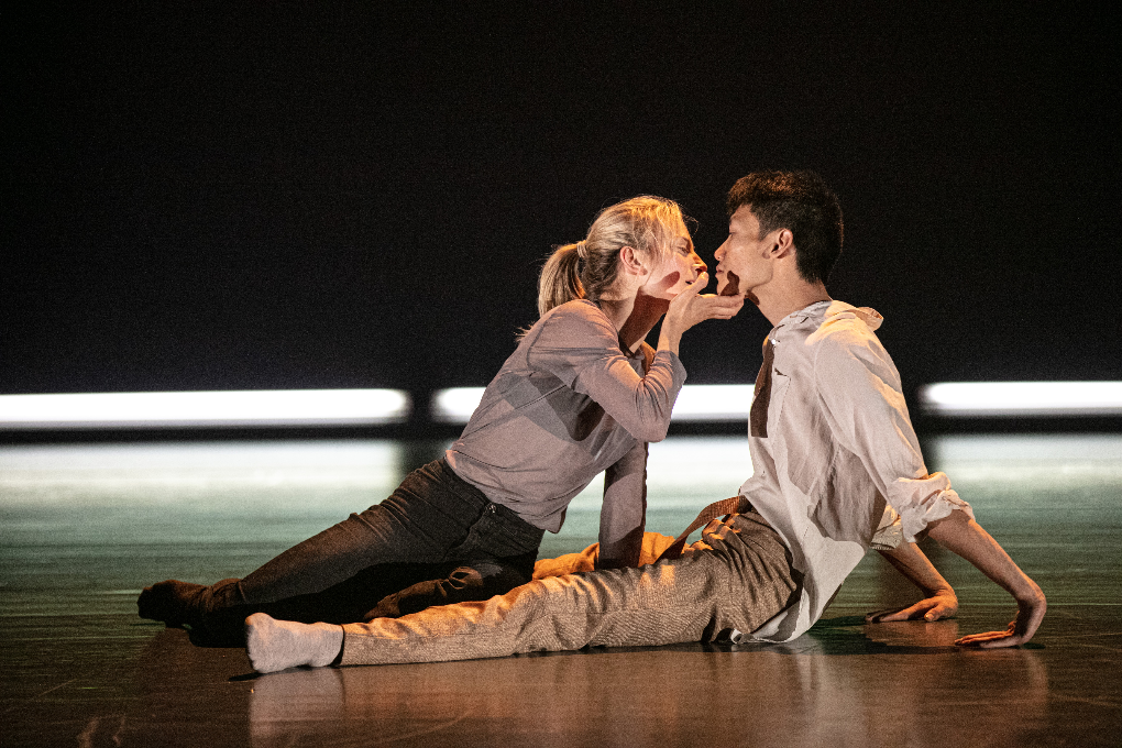Two dancers sitting on a stage in grey and beige clothing. The dancer on the left uses one hand to hold the face of the other dancer, leaning towards them. The backdrop is black with bright white lights along the floor.
