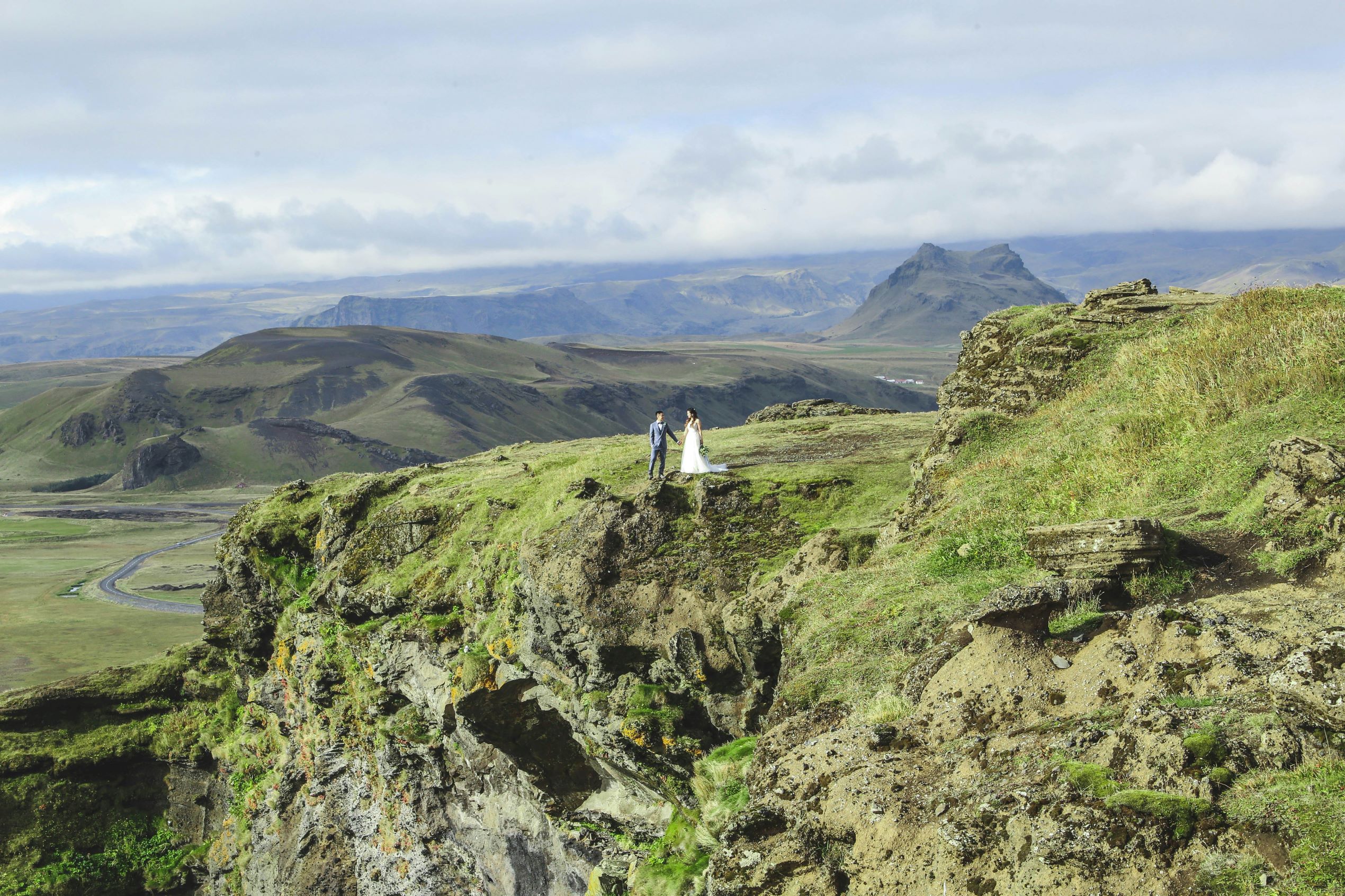 a couple dressed in wedding attire stand atop a green cliff, with impressive Icelandic mountain landscapes in the background