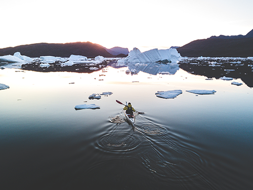 A person in a yellow jacket paddles a kayak towards some large chunks of iceberg in Greenland 