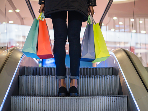A picture of the back of someone's legs making their way up the escalator in a shopping mall, holding four brightly coloured shopping bags