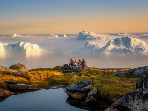 people atop a mountain in Greenland, looking at a dramatic view of icebergs and mountains