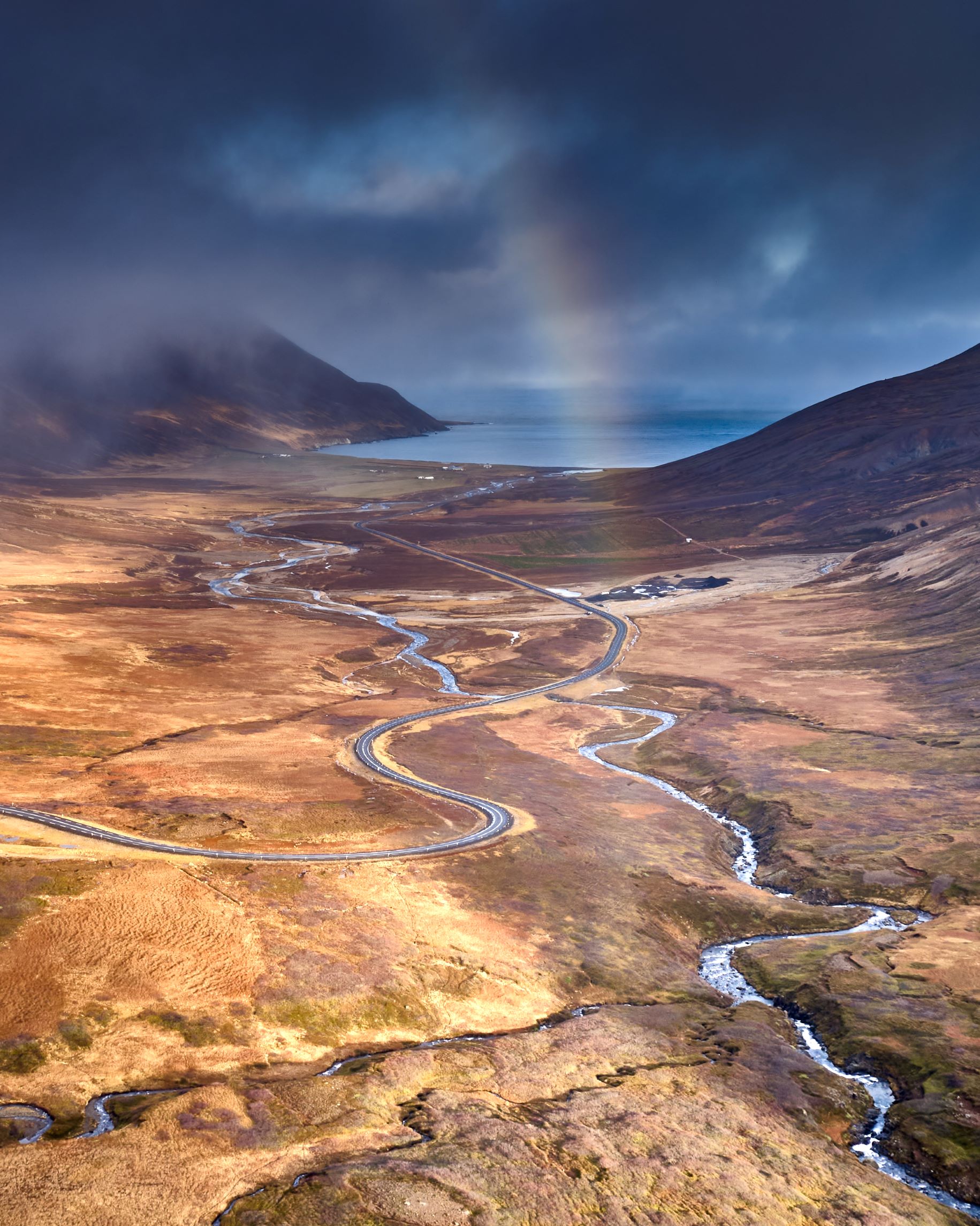 An elevated view of a road twisting down to a fjord edge, with a rainbow and cloudy skies overhead