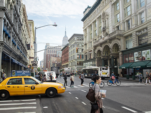A street level scene of New York, with a woman walking in the fore of the shot and a yellow cab to the left of frame and several people walking and cycling behind 