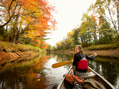 A young woman paddles in a canoe on water surrounded by trees covered in autumn leaves 