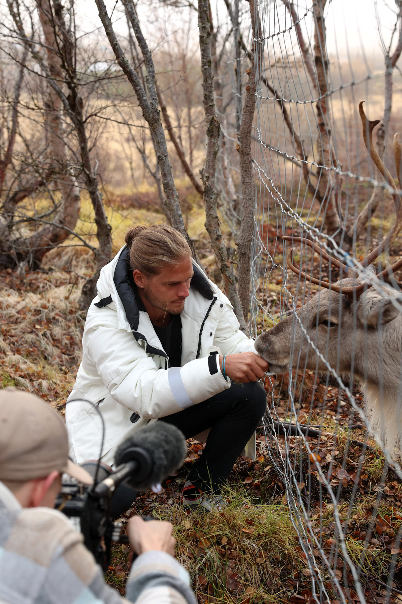 Rúrik Gíslason pets a reindeer at an enclosure while a cameraman looks on