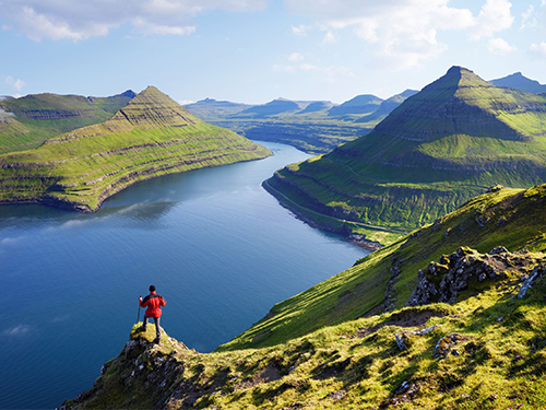 A man in a red rain jacket is pictured looking out over the mountainous landscapes of the Faroe Islands from atop a cliff edge