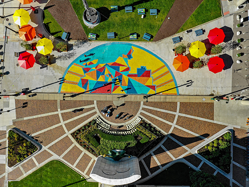 A birds eye view of a central square in Detroit, with people mulling around, bright umbrellas put up, and a beautiful street art piece on the pavement below 