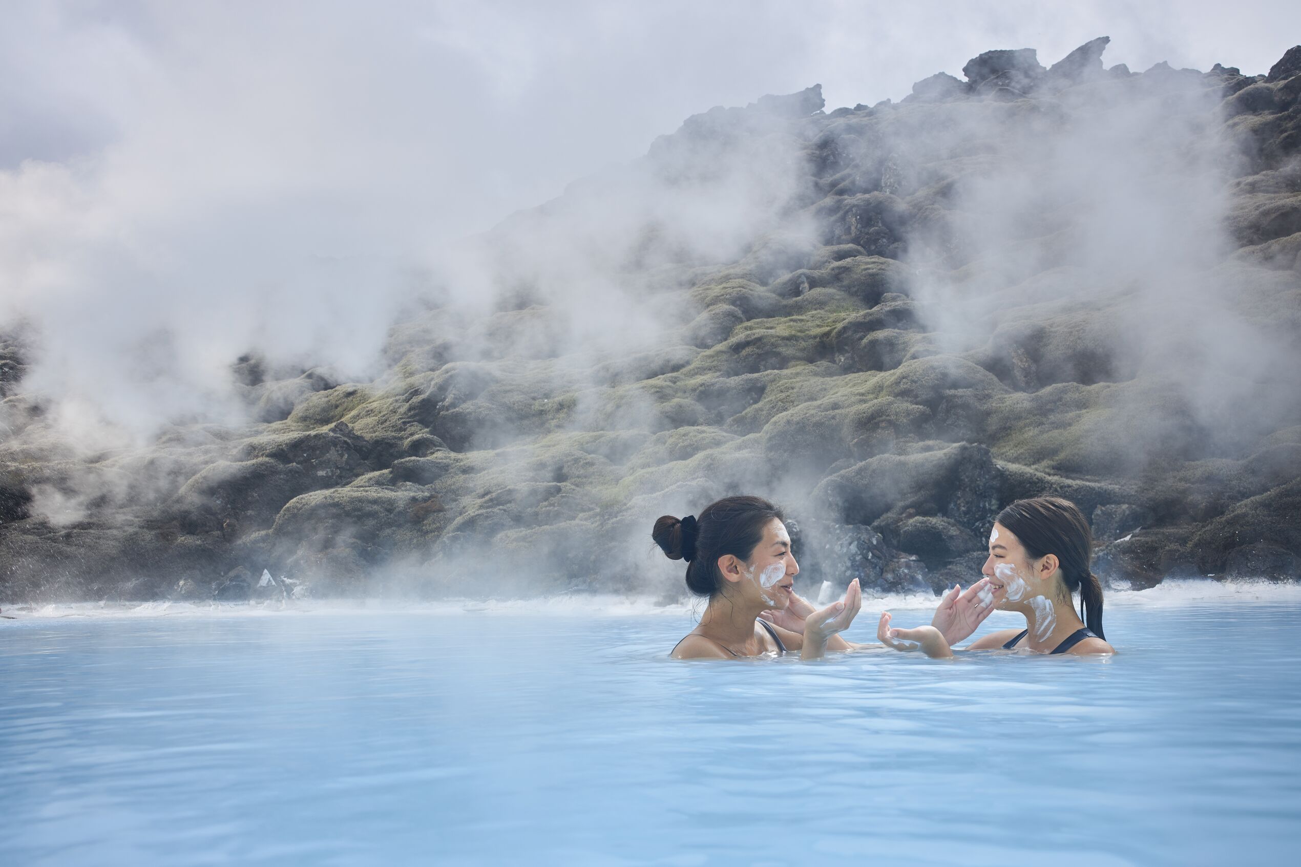 two women putting silica mud on each other faces the blue lagoon, Iceland.