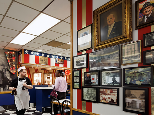 An interior of a restaurant in Detroit, with a server walking towards the camera holding several bottles of beer and people waiting to be served at the bar 