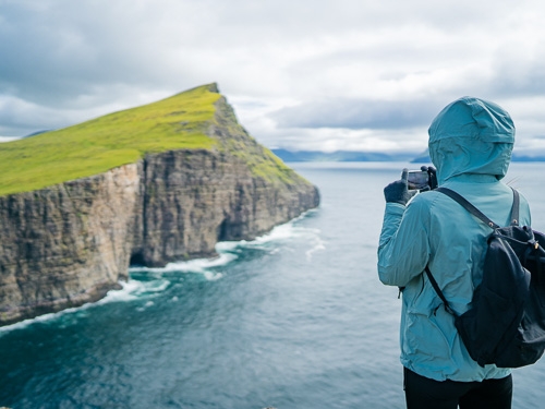 En person i en blå regnjakke står vendt mod et smukt kystlandskab med et kamera i hænderne. 