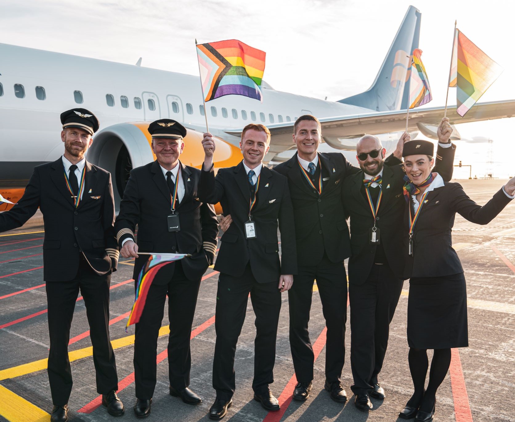 Icelandair's Pride flight 2022: 2 pilots and 4 cabin crew stand in front of Icelandair plane, waving rainbow flags.