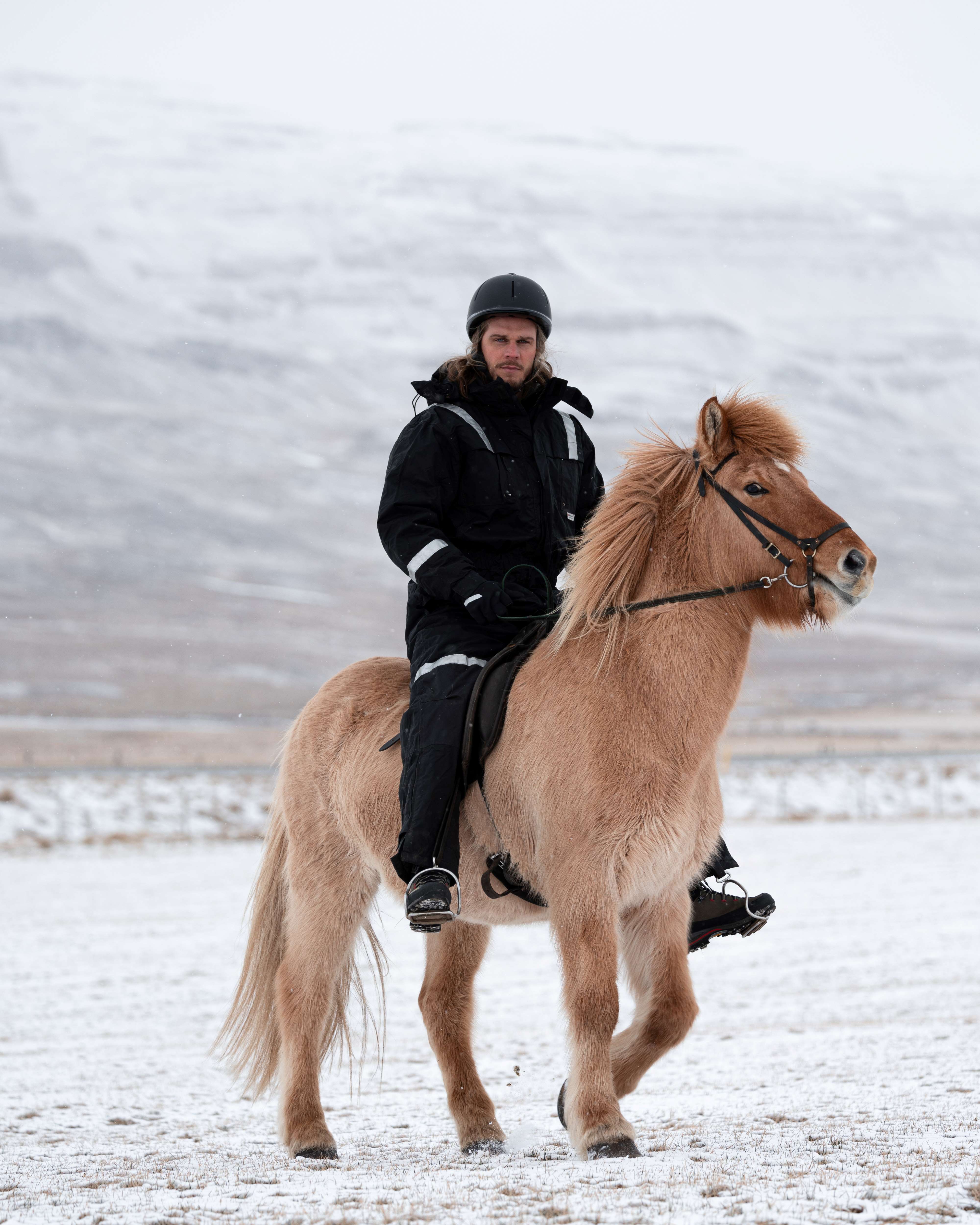 Rúrik Gíslason sits astride a sand-colored Icelandic horse, with moiuntains in the background.