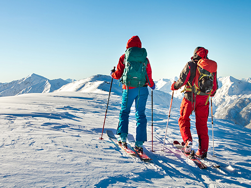 Two people moving up a mountain on skis while wearing colourful backpacks 