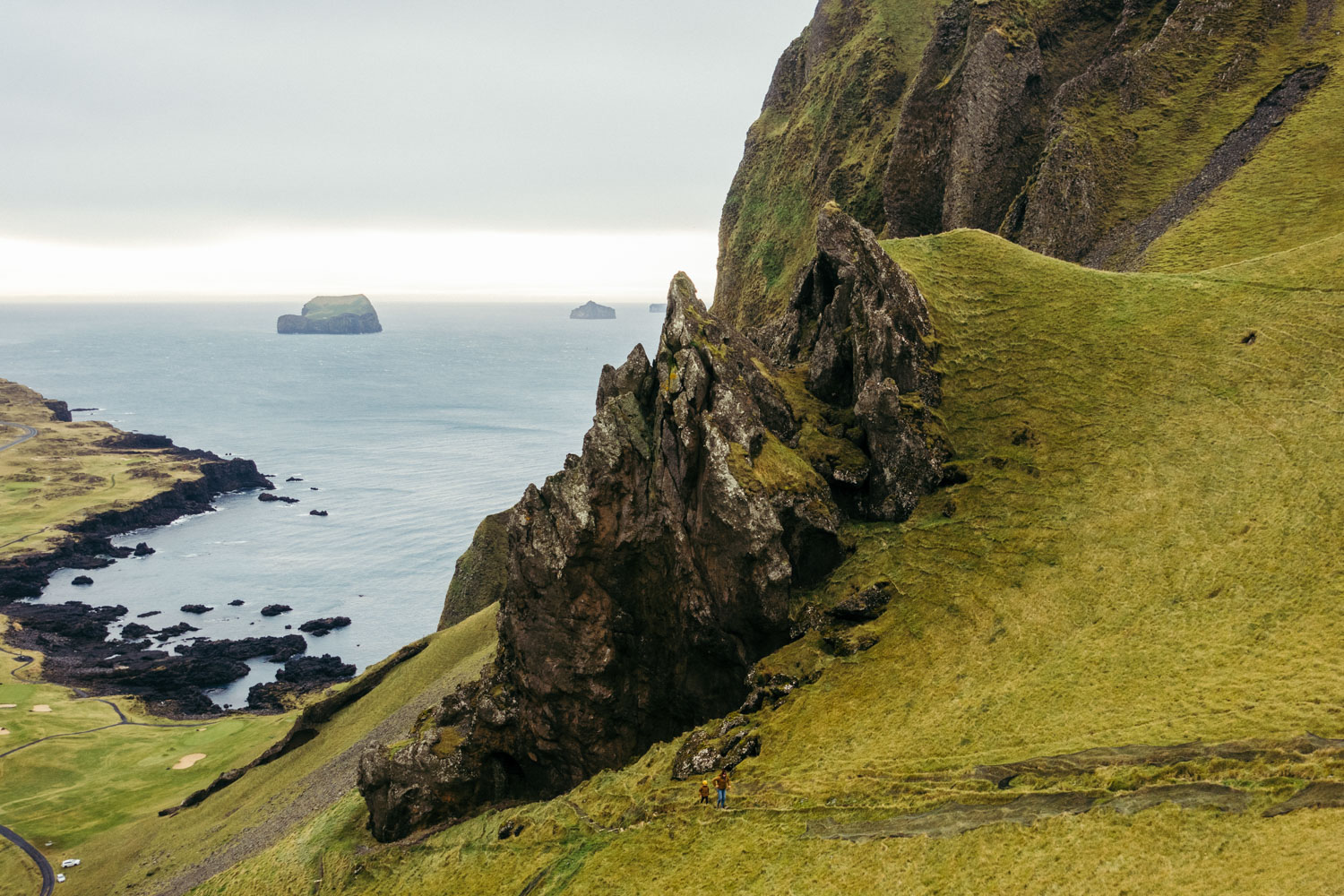 Image of tiny figures climbing a steep mountain track on a hill in Vestmannaeyjar, with ocean in background