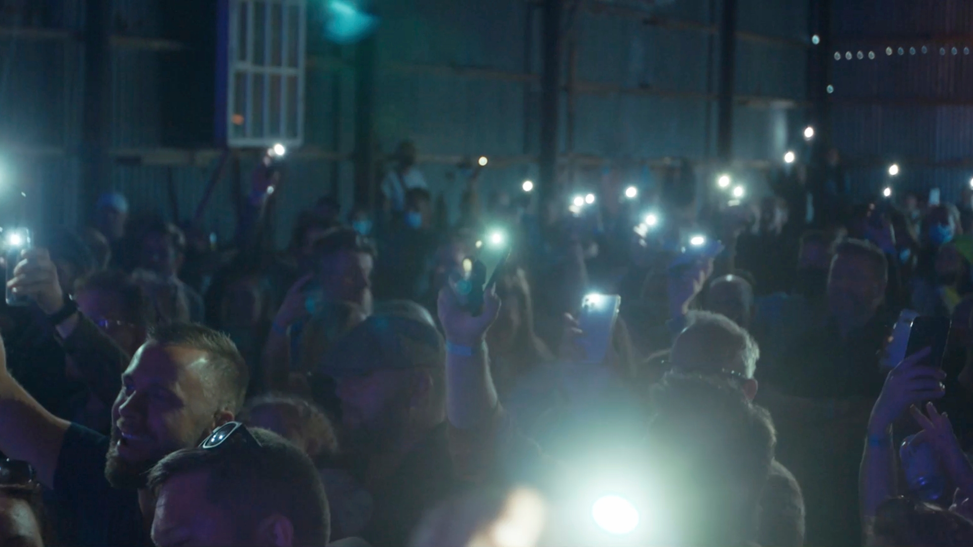 Inside the factory, a large crowd stands in a darkened room with their lit up phones held overhead, enjoying Braedlan music festival
