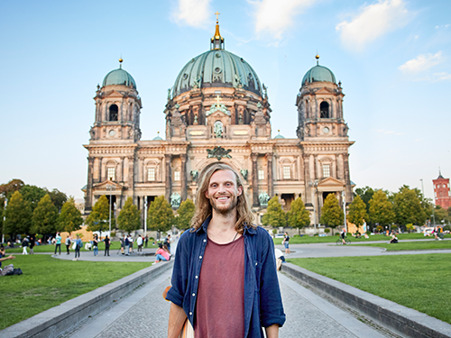A man with long blonde hair in center shot with the beautiful Berlin Cathedral Church in the background