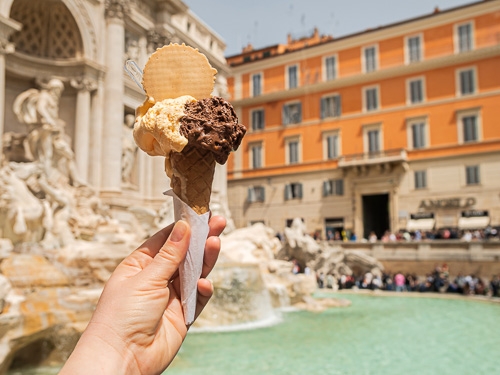 A hand holds a chocolate and vanilla ice cream cone up in front of the Trevi Fountain in Rome