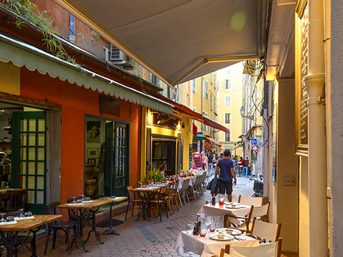 A view of one of the many narrow streets in Nice, France with cafes and restaurants on both sides of the street and people milling around 