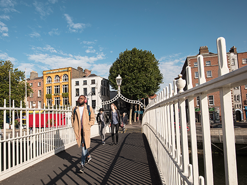 Three people walk across the Ha’Penny Bridge in Dublin during the day, with blue skies in the background 